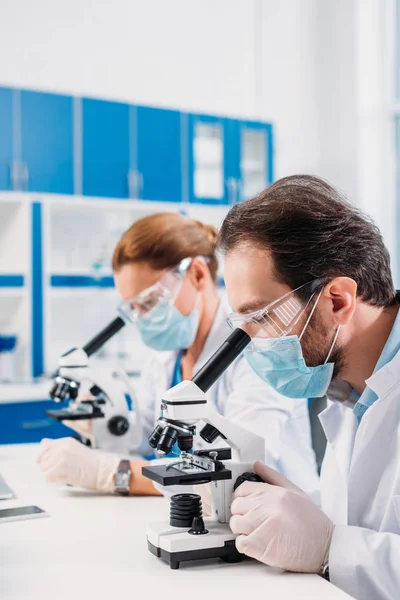 Enfoque selectivo de los científicos en máscaras médicas y gafas mirando a través de microscopios en regentes en el laboratorio - foto de stock