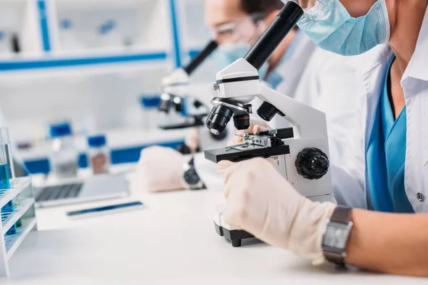 Selective focus of scientists in medical masks and goggles looking through microscopes on regents in lab — Stock Photo