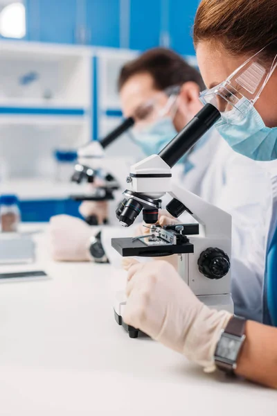 Enfoque selectivo de los científicos en máscaras médicas y gafas mirando a través de microscopios en regentes en el laboratorio - foto de stock