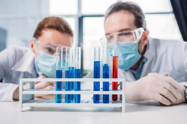 Enfoque selectivo de los investigadores científicos en googles y máscaras médicas mirando reactivos en tubos en laboratorio - foto de stock