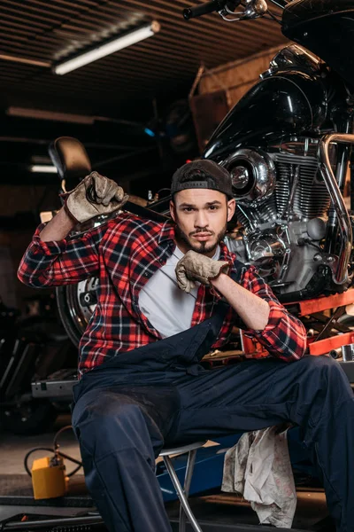 Atractivo trabajador de la estación de reparación de bicicletas con llave inglesa sentado delante de la motocicleta - foto de stock