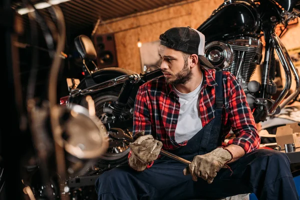 Bel lavoratore stazione di riparazione bici con chiave inglese seduto di fronte alla moto — Foto stock