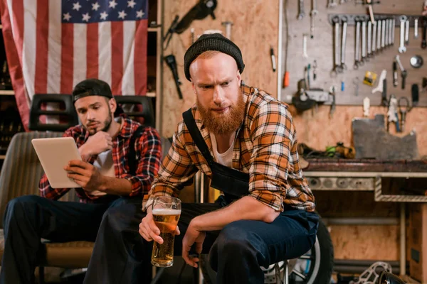 Mecánico guapo beber cerveza mientras que su colega el uso de tableta en el fondo en el garaje - foto de stock