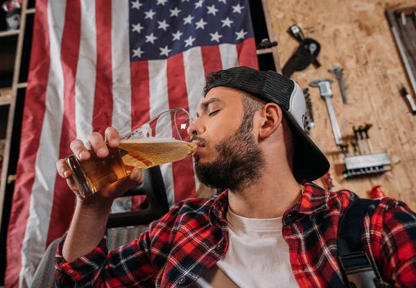 Trabajador de la estación de reparación guapo beber cerveza en el garaje con bandera EE.UU. colgando en la pared - foto de stock