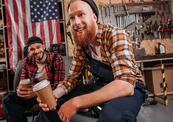 Happy mechanics drinking coffee from disposable cups on break at garage — Stock Photo