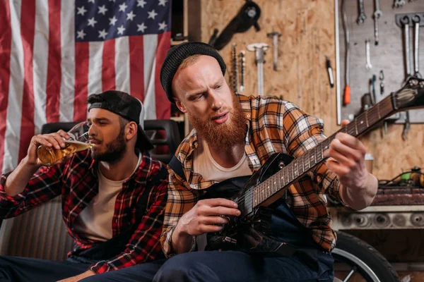 Mecánicos guapos tocando la guitarra y beber cerveza en el garaje - foto de stock