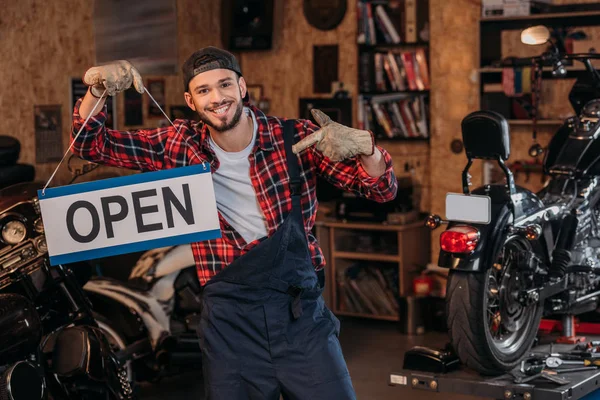 Feliz trabalhador da estação de reparo da bicicleta apontando para tabuleta aberta na garagem — Fotografia de Stock