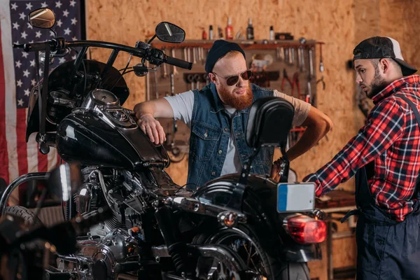 Joven trabajador de reparación hablando con el cliente serio cerca de la motocicleta en el garaje - foto de stock