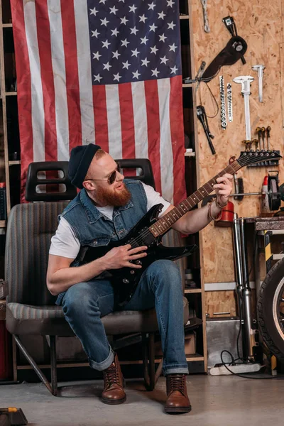 Красивый бородатый мужчина играет на электрогитаре в гараже с флагом США висит на стене — стоковое фото