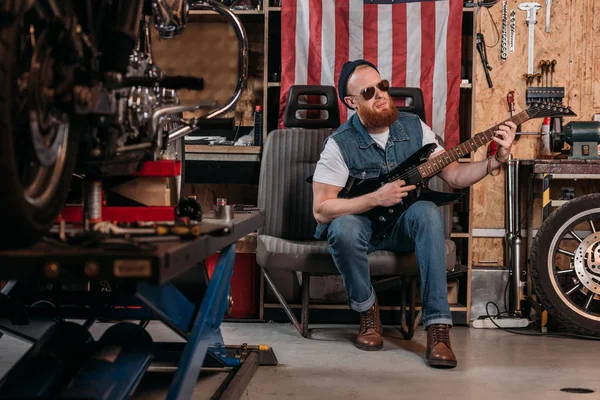 Barbudo hombre jugando guitarra eléctrica en el garaje con bandera de EE.UU. colgando en la pared - foto de stock