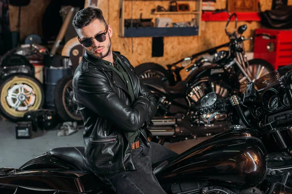 Schöner junger Mann mit Sonnenbrille und Lederjacke auf Fahrrad in Garage — Stockfoto