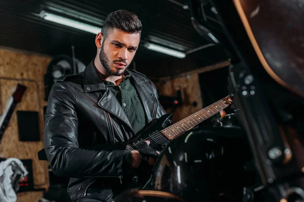 Schöner junger Mann spielt Gitarre, während er auf dem Fahrrad in der Garage sitzt — Stockfoto