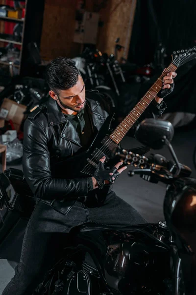 Handsome young man playing guitar while sitting on bike at garage — Stock Photo