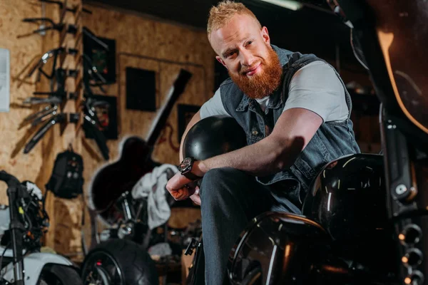 Jeune homme barbu assis sur un vélo avec casque au garage — Photo de stock