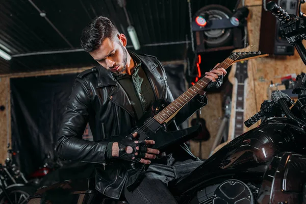 Schöner junger Mann spielt Gitarre, während er auf dem Fahrrad in der Garage sitzt — Stockfoto