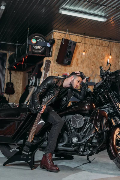 Beau jeune homme assis sur un vélo avec guitare électrique au garage — Photo de stock