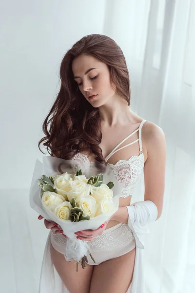 Femme chère en sous-vêtements avec bouquet de roses dans les mains debout à la fenêtre — Photo de stock