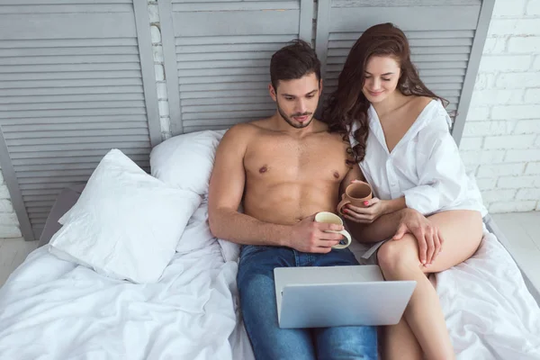 Молода пара з чашками кави використовує ноутбук під час відпочинку на ліжку вдома — стокове фото