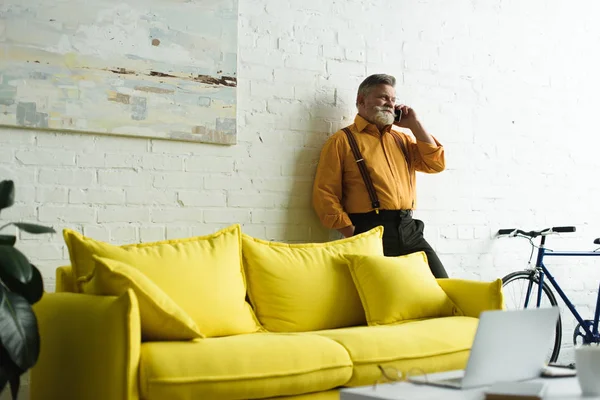 Hombre mayor barbudo con estilo hablando por teléfono inteligente y mirando hacia fuera en casa - foto de stock