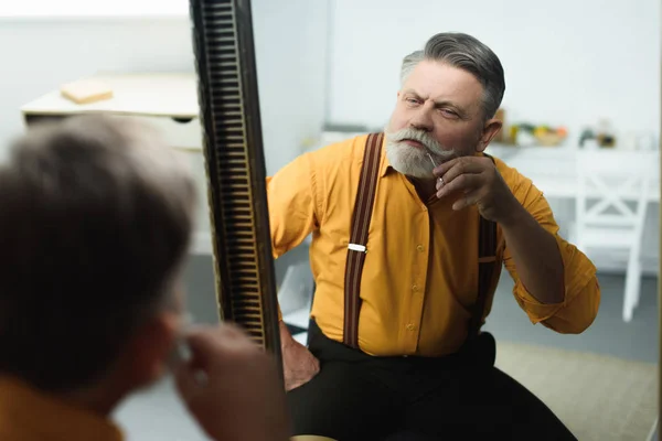 Guapo barbudo hombre mayor recortar bigote con tijeras y mirando al espejo - foto de stock