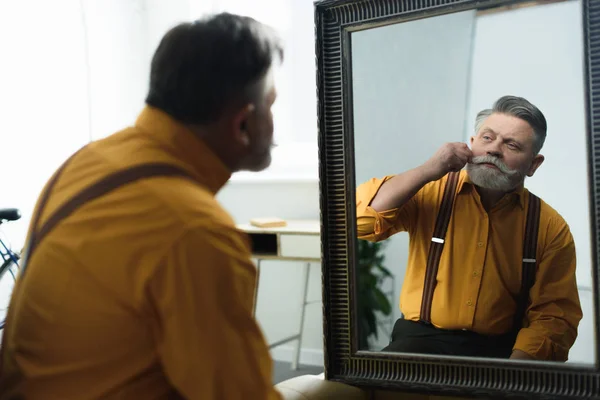 Красивый бородатый мужчина, регулирующий усы и смотрящий в зеркало — стоковое фото