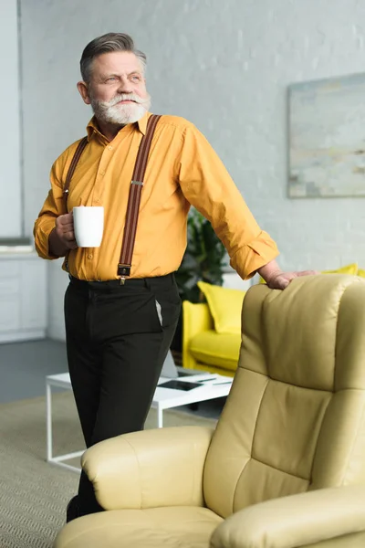 Hombre mayor sosteniendo la taza de café y mirando hacia otro lado mientras está de pie cerca del sillón en casa - foto de stock