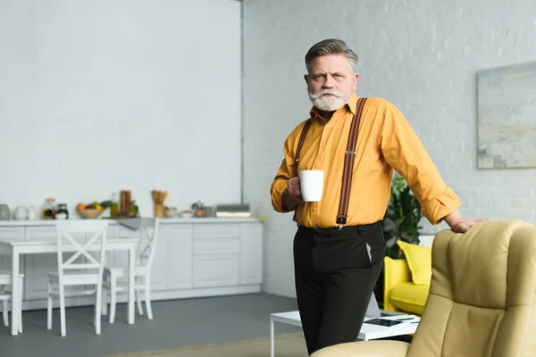 Homem sênior barbudo elegante segurando xícara de café e olhando para a câmera em casa — Fotografia de Stock