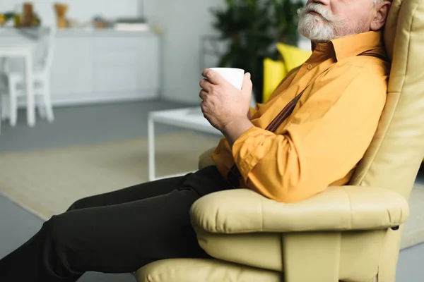 Schnappschuss eines bärtigen Seniors, der eine Tasse in der Hand hält und zu Hause auf einem Sessel sitzt — Stockfoto