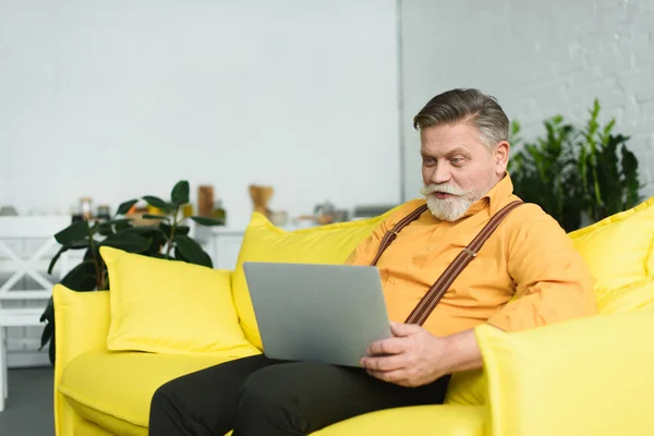 Homme âgé barbu souriant utilisant un ordinateur portable tout en étant assis sur le canapé à la maison — Photo de stock