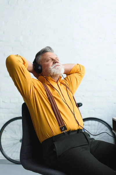 Sorridente barbudo idoso em fones de ouvido ouvindo música enquanto sentado com as mãos atrás da cabeça em casa — Fotografia de Stock