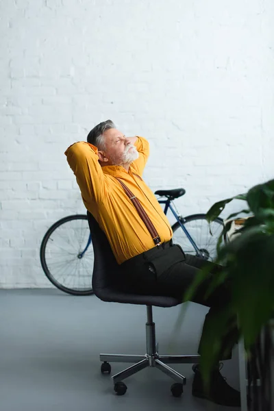 Seitenansicht eines bärtigen älteren Mannes, der mit den Händen hinter dem Kopf auf einem Stuhl sitzt — Stockfoto