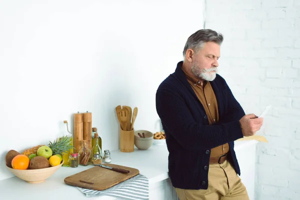 Bearded senior man using smartphone while standing at kitchen — Stock Photo