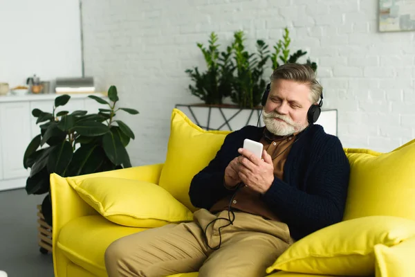 Uomo anziano barbuto sorridente in cuffia seduto sul divano e ascoltare musica con smartphone — Foto stock