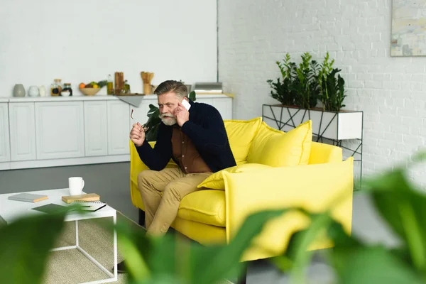 Selektiver Fokus eines älteren Mannes, der mit dem Smartphone spricht und wegschaut, während er zu Hause auf dem Sofa sitzt — Stockfoto