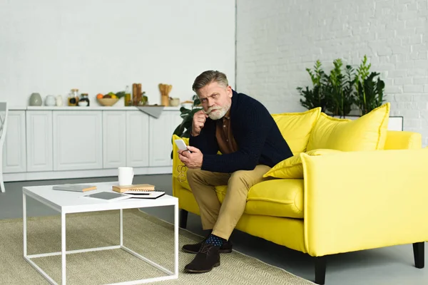 Schöner älterer Mann mit Smartphone und Blick in die Kamera, während er zu Hause auf dem Sofa sitzt — Stockfoto