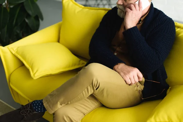 Cropped shot of senior man holding eyeglasses and talking by smartphone while sitting on sofa — Stock Photo