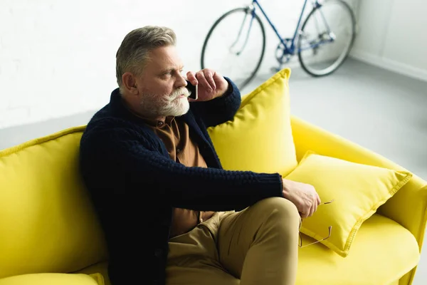 Bearded senior man talking by smartphone and looking away while sitting on sofa at home — Stock Photo