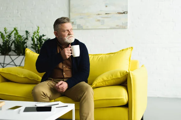 Pensativo hombre mayor sosteniendo la taza de té mientras está sentado en el sofá amarillo y mirando hacia otro lado en casa - foto de stock