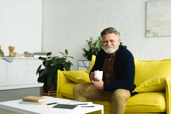 Barbudo hombre mayor sosteniendo la taza de café y sonriendo a la cámara en casa - foto de stock