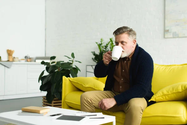 Senior man sitting on yellow sofa and drinking tea at home — Stock Photo