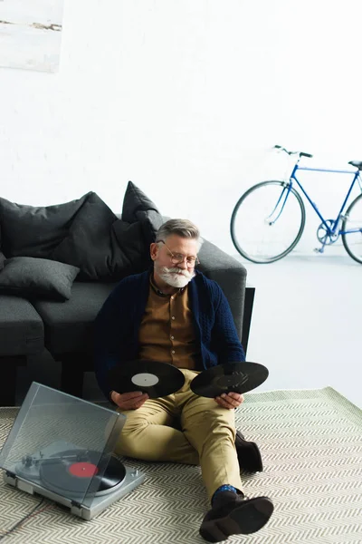 Hochwinkelaufnahme eines bärtigen älteren Mannes mit Brille, der Schallplatten hält, während er zu Hause auf dem Teppich sitzt — Stockfoto