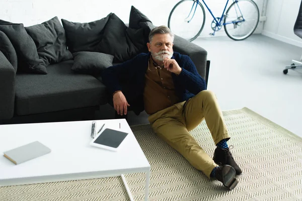Високий кут зору красивий стильний старший чоловік тримає окуляри, сидячи на килимі вдома — стокове фото