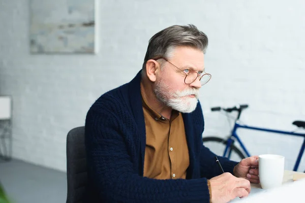 Thoughtful senior man in eyeglasses holding cup and taking notes at home — Stock Photo