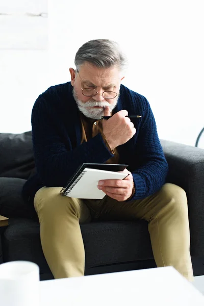 Serious senior man in eyeglasses holding pen and notebook while sitting on sofa — Stock Photo