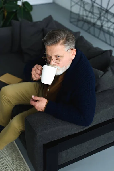 Vue grand angle de bel homme dans des lunettes buvant de la tasse et regardant loin tout en étant assis sur le canapé à la maison — Photo de stock