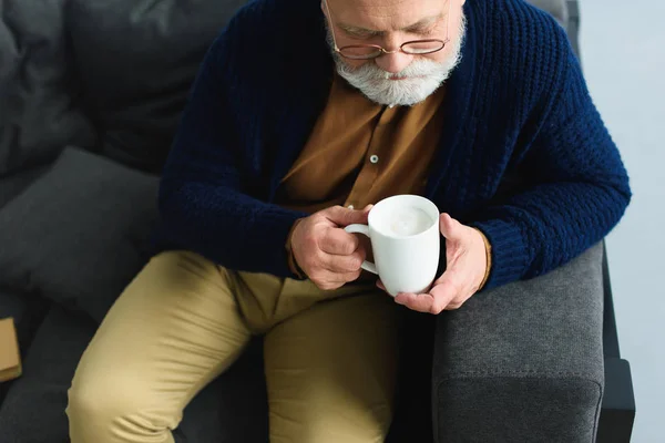 Tiro recortado de hombre mayor barbudo en gafas con taza mientras está sentado en el sofá - foto de stock