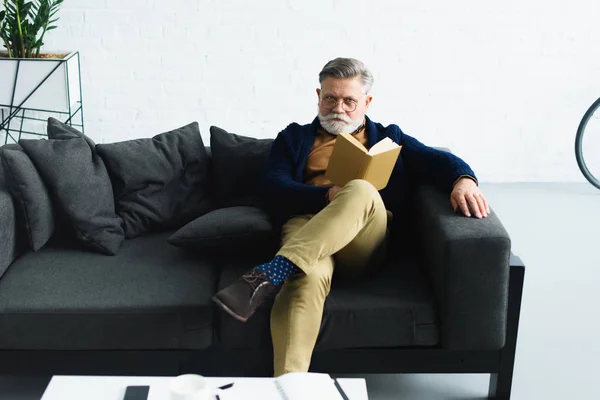 Stilvoller bärtiger Mann mit Brille, Buch in der Hand und in die Kamera blickend, während er zu Hause auf dem Sofa sitzt — Stockfoto