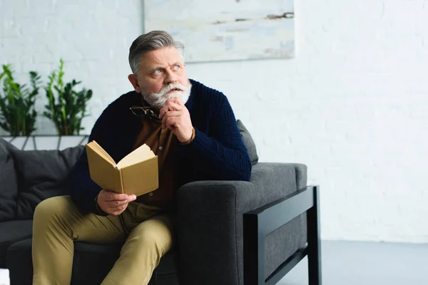 Pensive stylish senior man holding book and looking away at home — Stock Photo