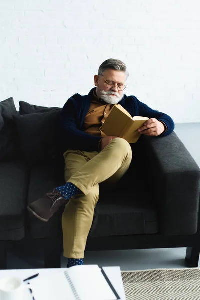 Стильний бородатий чоловік читає книгу на дивані вдома — стокове фото