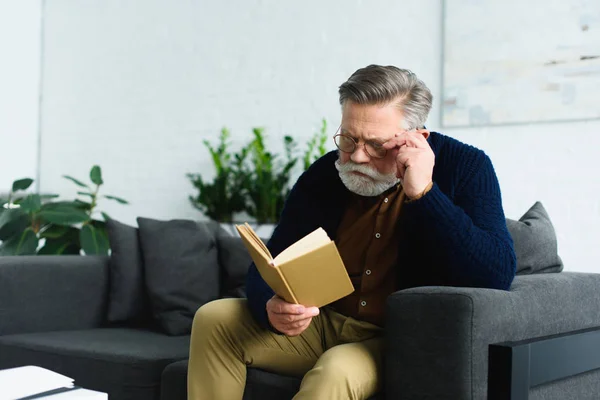 Stylish senior man in eyeglasses sitting on sofa and reading book at home — Stock Photo